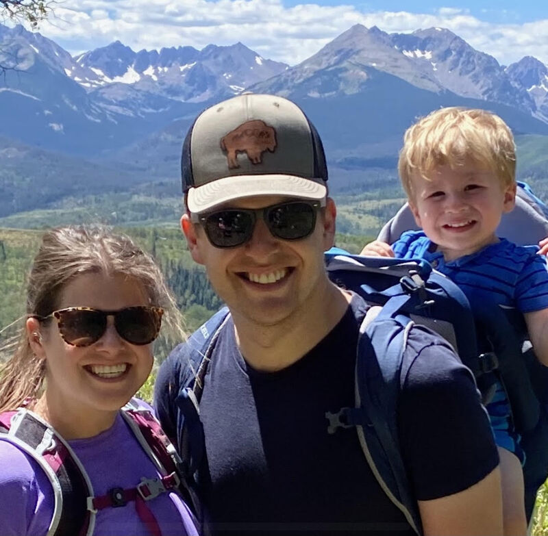 The image shows a family of three posing for a photo in front of a scenic mountain landscape. The father is wearing a hat and sunglasses, and he has a young child in a backpack carrier. The mother is standing next to him, also wearing sunglasses. They are all smiling and appear to be enjoying their time outdoors. The background features lush greenery and towering mountains under a bright sky.